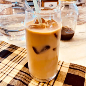 A tall glass of creamy brown delicious looking iced cold brew coffee. You can see the cold brew coffee in the background. The coffee is served in a tall glass with multiple ice cubes and a straw.