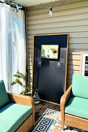 DIY home decor dor the front porch. A large chalkboard for outdoors sitting on a deck with a chalk drawn shelf and a real image hanging from the chalkboard. It is flanked by two turquoise cushioned chairs, a white curtain and plants.