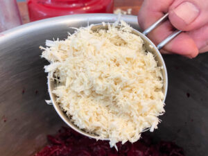 Adding a measuring cup full of freshly grate horseradish to a large stainless steel pot for making beet relish.