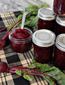 Several jars of homemade beet relish with horseradish are stacked on the right. There is a open jar of relisj on the left and two freshly cooked garden beets for decoration. Everything is sitting on a plaid kitchen towel.