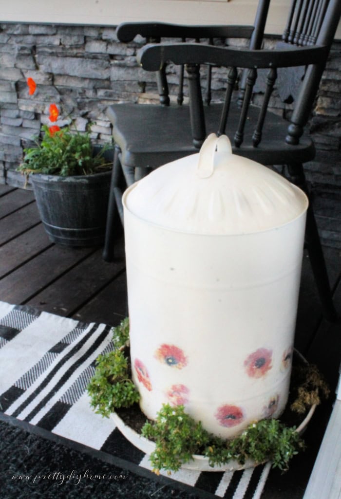 Early fall porch decor showing a refinished DIY chicken feeder in cream with poppies, and a poppy plant. There is also a black chair, plaid black and white carpet and a black rug.