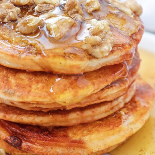 A stack of five fluffy and delicious pumpkin pancakes sitting on a whhite plate. The are drizzled in lots of maple syrup and topped with walnuts.