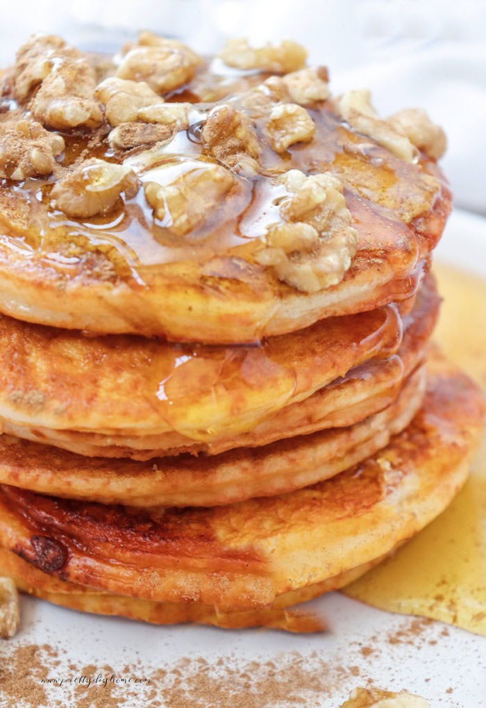 A stack of five fluffy and delicious pumpkin pancakes sitting on a whhite plate. The are drizzled in lots of maple syrup and topped with walnuts.