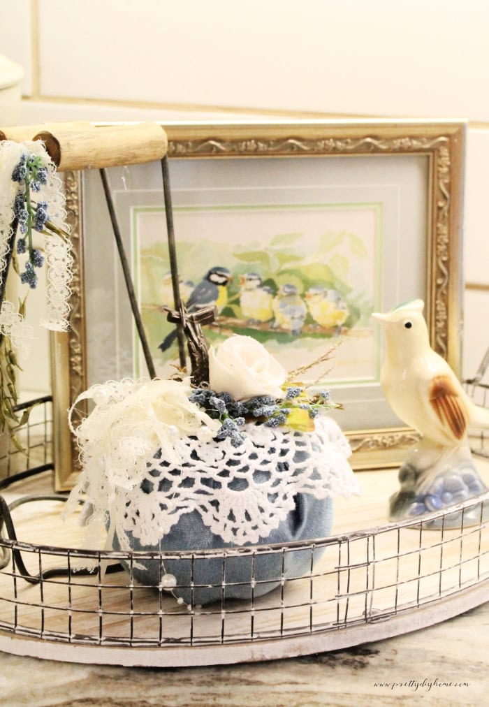 A Dollar Tree pumpkin for fall decorated with a small vintage doily. This easy fall craft shows the blue of the pumpkin bottom and has assorted flowers are ribbons on the top. The vintage doily pumpkin is sitting on a kitchen tray with various vintage items, a vintage crock, a vintage blue bird, and a vintage blue bird picture in the background.