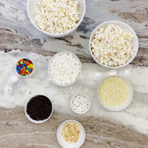Halloween popcorn ingredients in various small white bowls, There is popcorn, marshmallowed M&Ms melted white chocolate, candy eyeballs and peanuts.
