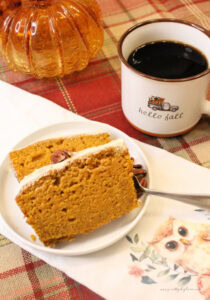 Two slices of homemade pumpkin bread with sour cream glaze, sitting on a white plate. The plate is sitting on a tea towel with an owl decoration, and the tea towel is sitting on a plaid table cloth. There is a cup of coffee in the background.