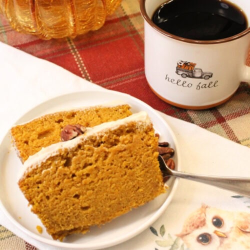 Two slices of homemade pumpkin bread with sour cream glaze, sitting on a white plate. The plate is sitting on a tea towel with an owl decoration, and the tea towel is sitting on a plaid table cloth. There is a cup of coffee in the background.