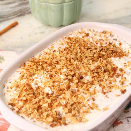A large pumpkin crisp with whipped cream and granola. The pumpkin dessert is sitting on a fall tea towel with pink and orange pumpkins and their is a sage ceramic pumpkin in the background.