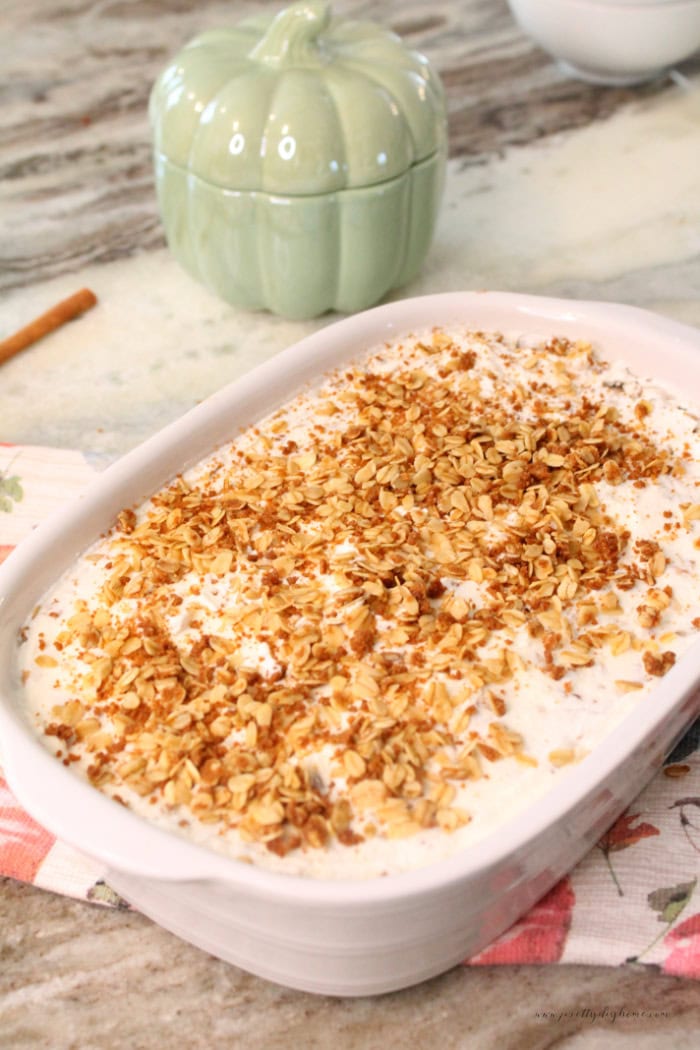 A large pumpkin crisp with whipped cream and granola. The pumpkin dessert is sitting on a fall tea towel with pink and orange pumpkins and their is a sage ceramic pumpkin in the background.