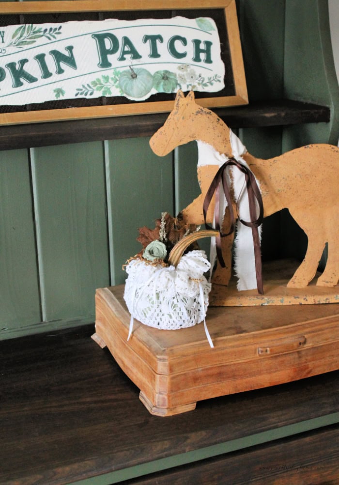 A Dollar Tree pumpki decorated with a large lace doily, thats tied with ribbon. The pumpkin has a real stem and is decorated with a rolled ribbon flower, and leaves. The pumpkin is white with a gold stem brown leaves and green flower and it sitting on a golden color vintage box withj a vintage metal horse in the same color standing behind it.