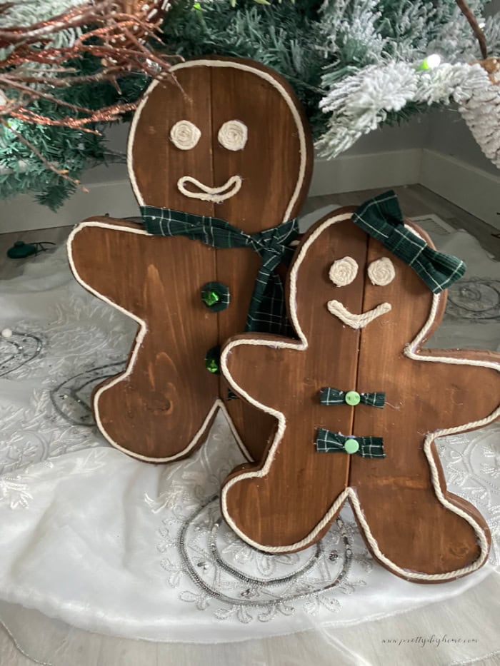 DIY wooden gingerbread man and woman decorated with macrame cord and green tartan accessories, displayed under a Christmas tree.