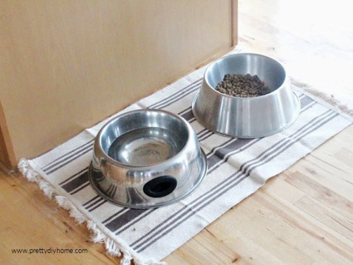 A DIY pet feeding mat before image showing dog bowls sitting on a small doormat on a hardwood floor.
