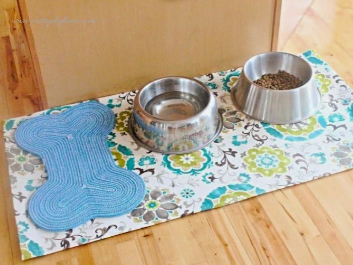 A large DIY pet feeding mat with two stainless steel dog bowls and a blue bone placemat on a hardwood floor.