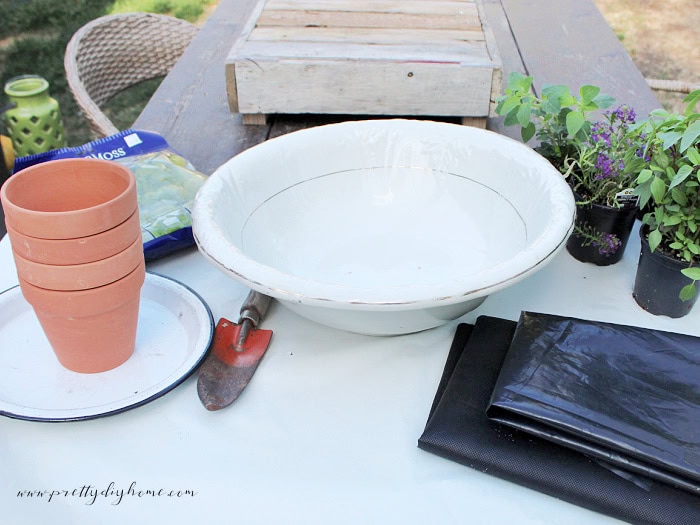 Supplies for creating a vintage container herb garden including folded fabric and plastic, herb plants, terracotta pots, moss, a porcelain bowl, and a vintage bulb shovel.