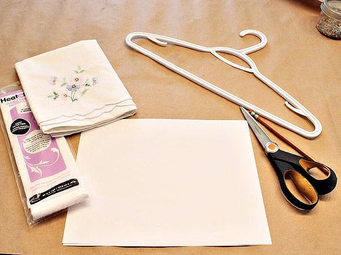 The supplies for making DIY hanger covers from repurposed vintage linens including a vintage embroidered pillowcase, hanger, interfacing, scissors, and pattern paper.