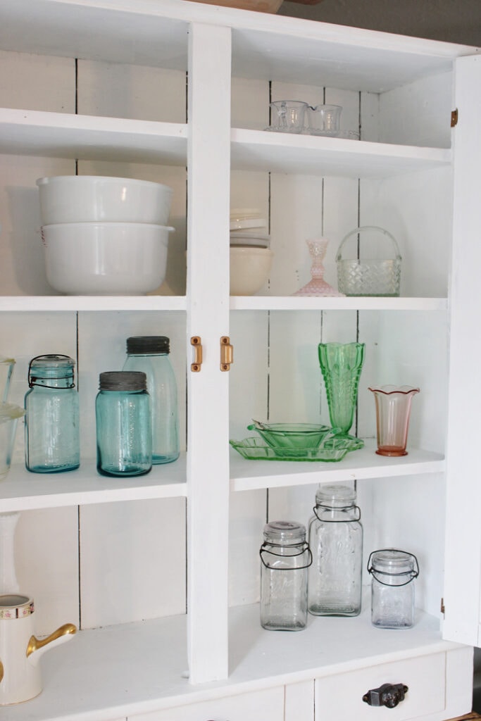 White painted vintage cabinet interior with open shelves styled using vintage dishes, green glassware, and cottage kitchen decor.
