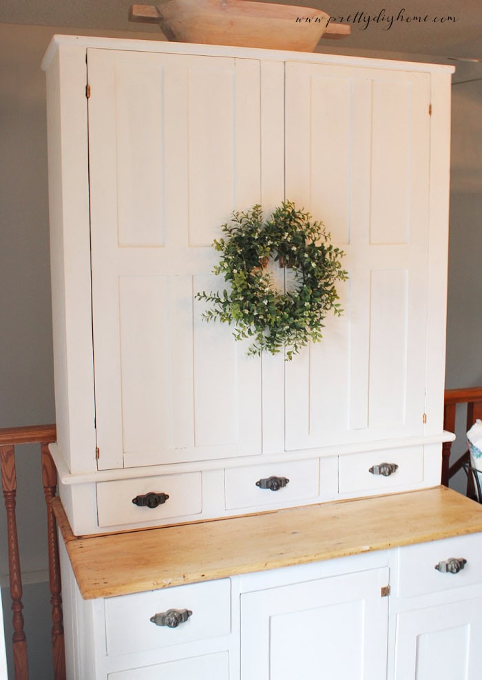 White vintage cabinet with closed doors, black hardware, and a decorative wreath styled in a cozy cottage dining room.
