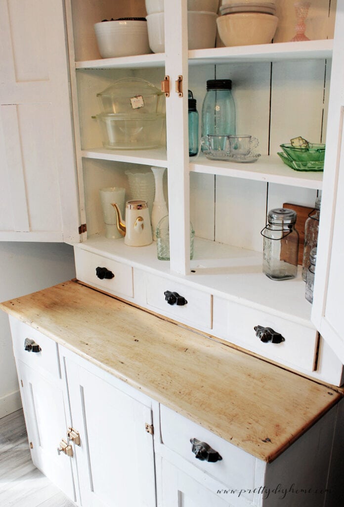 White vintage cabinet with open shelves and a natural wood countertop styled with vintage glassware and cottage kitchen decor.