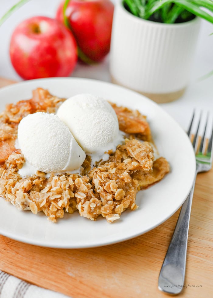 Apple crisp with vanilla ice cream served on a white plate with fresh apples and a potted plant in the background.