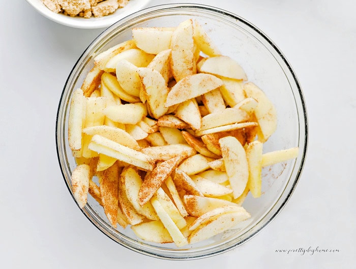 Fresh apple slices coated with cinnamon and sugar in a clear glass mixing bowl on a white marble countertop.