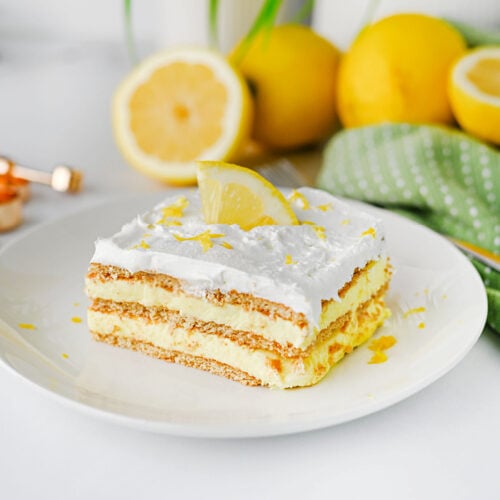 Easy lemon icebox cake topped with whipped cream, fresh lemon, and lemon zest. The creamy lemon dessert is served on a white plate with fresh lemons in the background.