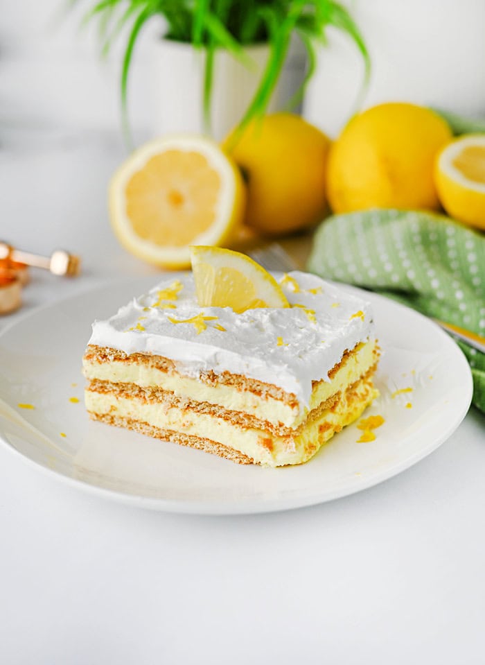 Easy lemon icebox cake topped with whipped cream, fresh lemon, and lemon zest. The creamy lemon dessert is served on a white plate with fresh lemons in the background.