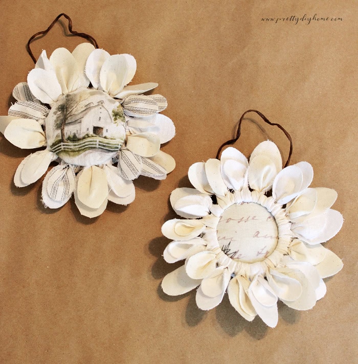 Two fabric flowers showing different center styles, one with a raised barn center glued on top and one with a flatter vintage letter center inserted into the ring.