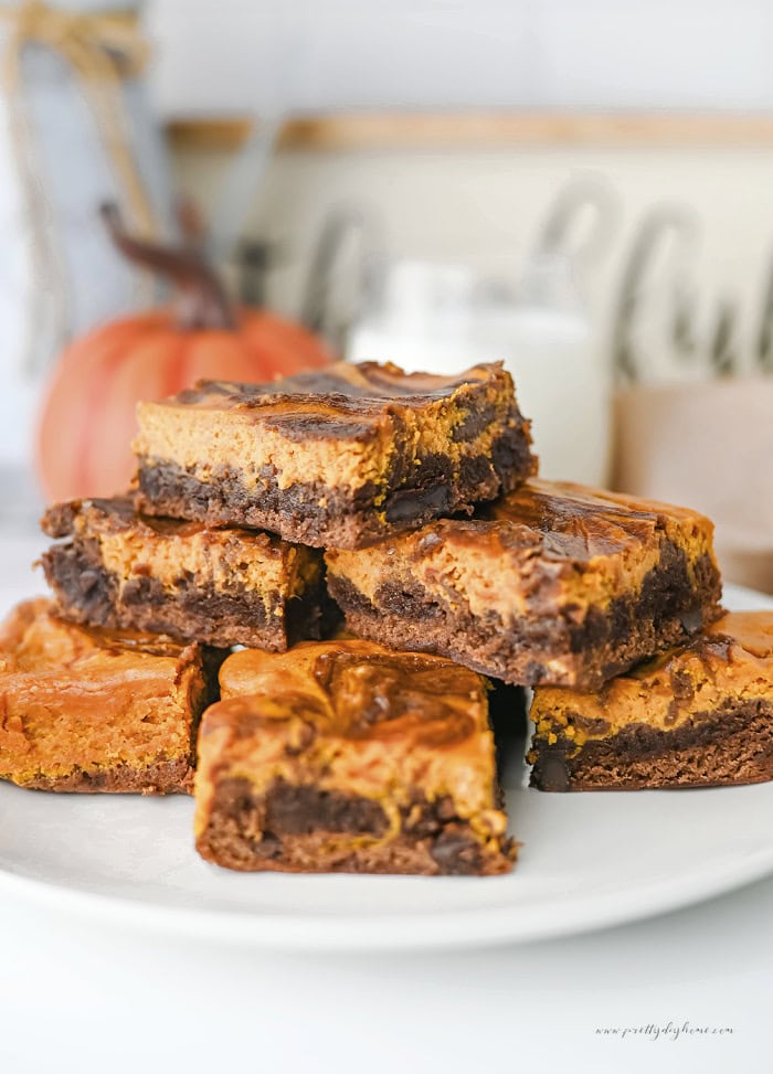 Stack of fudgy pumpkin brownies with a pumpkin cream cheese swirl and chocolate chips on a white serving plate with a pumpkin in the background.