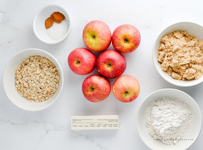 Ingredients for an old fashioned apple crisp including apples, oats, flour, brown sugar, butter, cinnamon, and sugar arranged in white bowls on a white marble countertop.