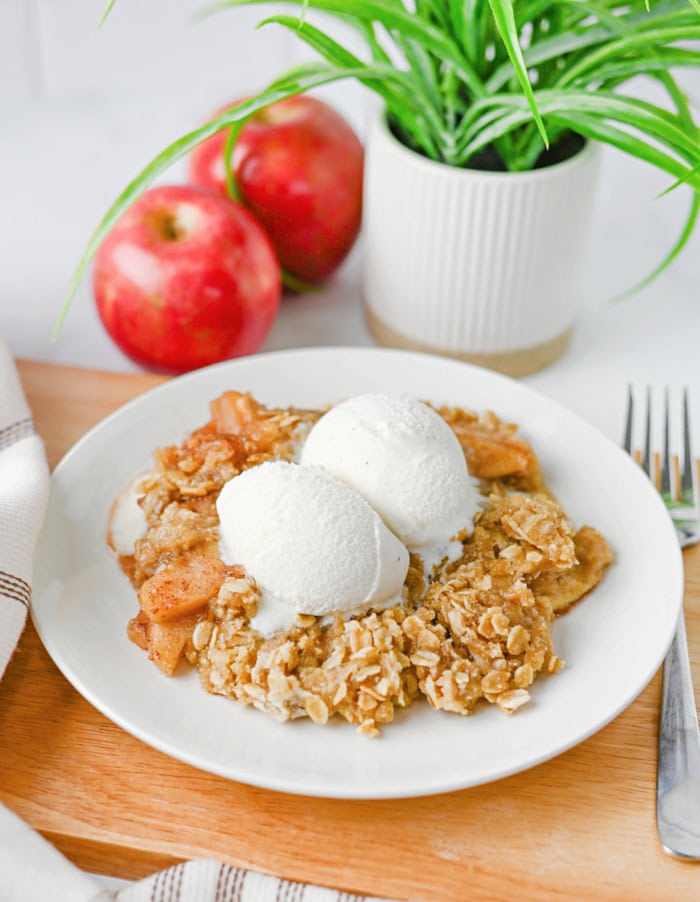 Old fashioned apple crisp with oat topping served on a white plate with two scoops of vanilla ice cream and fresh apples in the background.