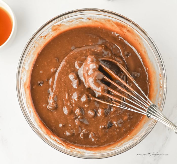 Chocolate chips being folded into pumpkin brownie batter with a whisk in a glass mixing bowl on a white marble countertop.