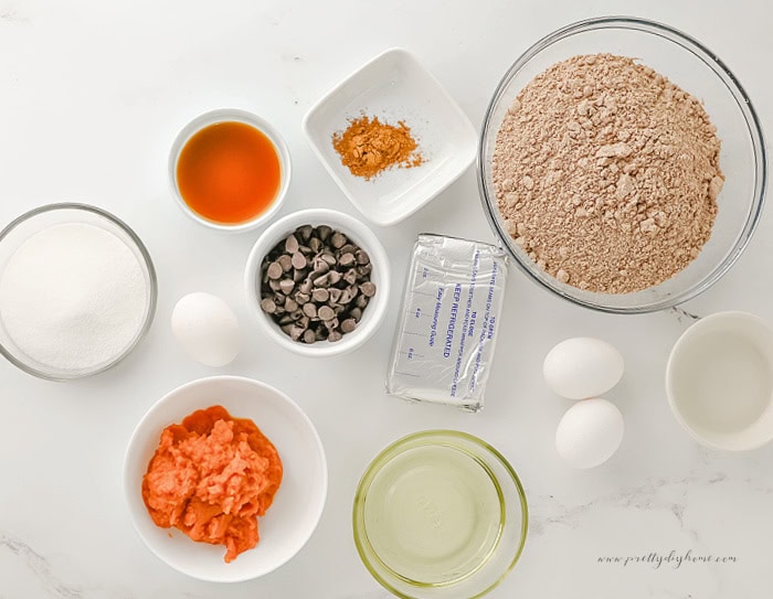 Ingredients for easy pumpkin brownies arranged in white ceramic and clear glass bowls on a white marble countertop, including brownie mix, pumpkin puree, cream cheese, chocolate chips, eggs, vegetable oil, sugar, vanilla extract, and pumpkin spice.