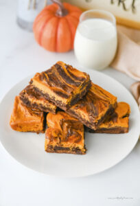Pumpkin brownies with a pumpkin cream cheese swirl served on a white plate with a glass of milk and a pumpkin in the background.