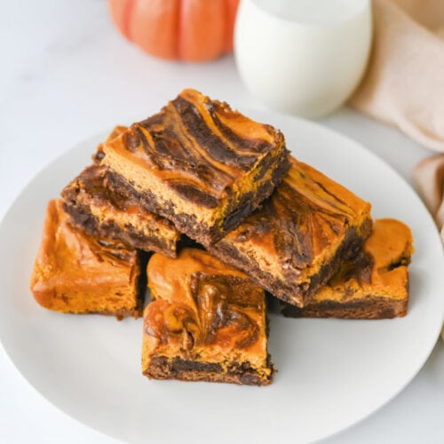 Pumpkin brownies with a pumpkin cream cheese swirl served on a white plate with a glass of milk and a pumpkin in the background.