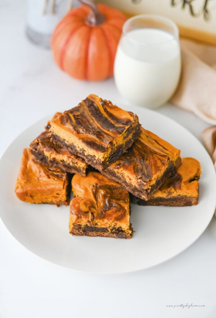 Pumpkin brownies with a pumpkin cream cheese swirl served on a white plate with a glass of milk and a pumpkin in the background.