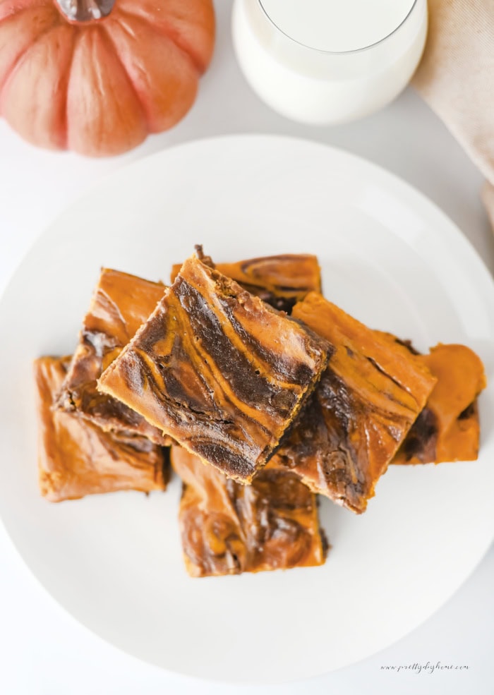 Pumpkin swirl brownies stacked on a white plate with a glass of milk and a pumpkin in the background.