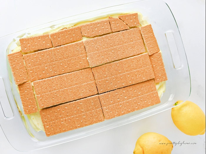 Second layer of graham crackers added to a lemon icebox cake. The cake is in a clear glass pan, and bits of the lemon filling peek out from underneath the graham crackers.