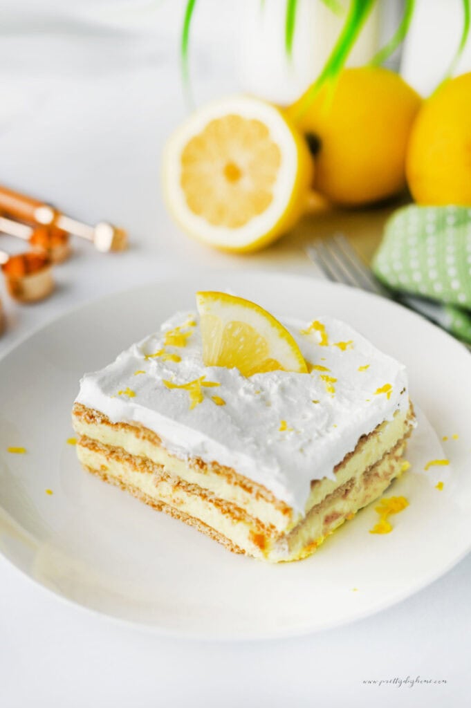A close up slice of lemon icebox cake topped with whipped cream, lemon zest, and a fresh lemon slice. The creamy lemon filling and graham cracker layers are clearly visible.