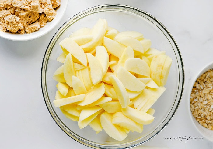 Freshly peeled and sliced apples in a clear glass mixing bowl on a white marble countertop with bowls of oats and brown sugar nearby.