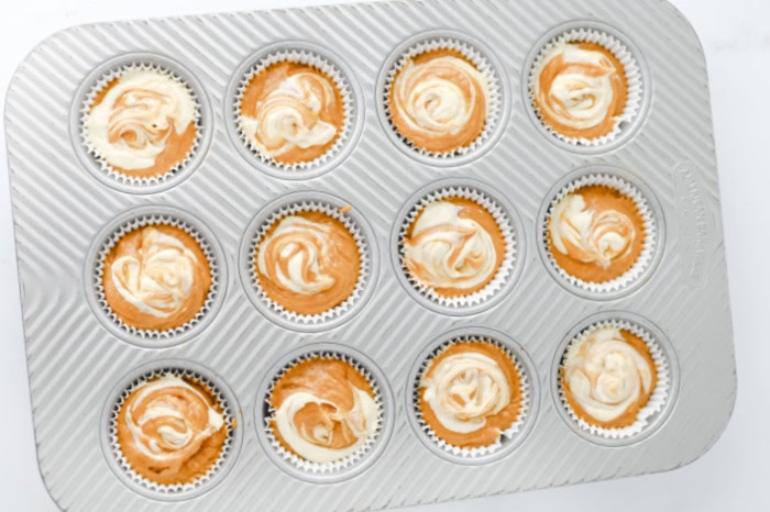 A muffin baking tray will with pumpkin batter and cheesecake filling thats been swirled.