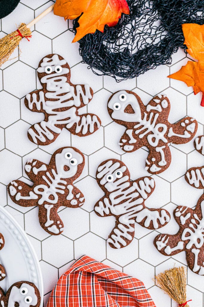 Several chocolate sugar cookies shaped like people are decorated with white icing and candy eyes to resemble playful skeletons and mummies.