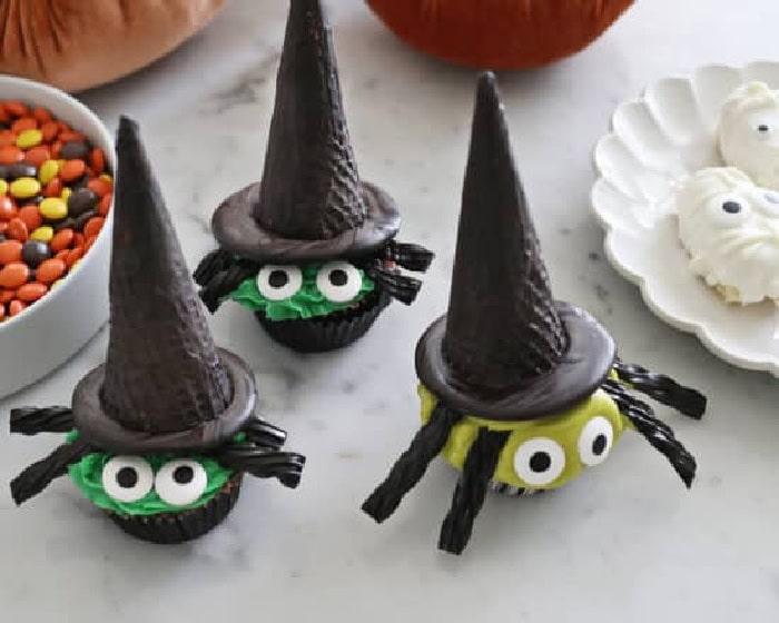 Three green witch cupcakes are decorated with candy eyes, black licorice hair, and tall chocolate cone hats on a marble counter.