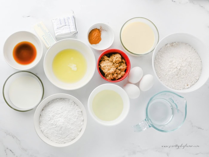 Ingredients for cinnamon roll poke cake arranged on a white counter, including cake mix, eggs, oil, cinnamon, brown sugar, butter, cream cheese, powdered sugar, vanilla, and sweetened condensed milk.