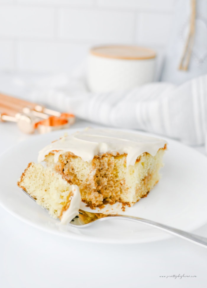 Slice of cinnamon roll poke cake topped with thick cream cheese frosting, with a forkful showing the soft vanilla cake and cinnamon brown sugar filling.