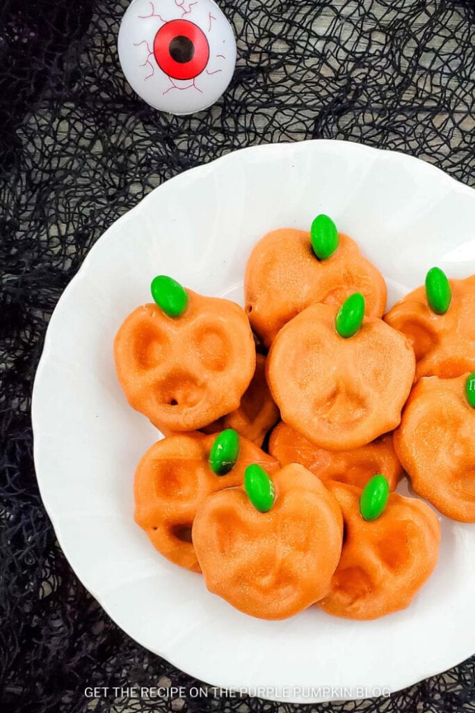 A plate of orange candy coated pumpkin pretzels with green candy stems on a white plate.