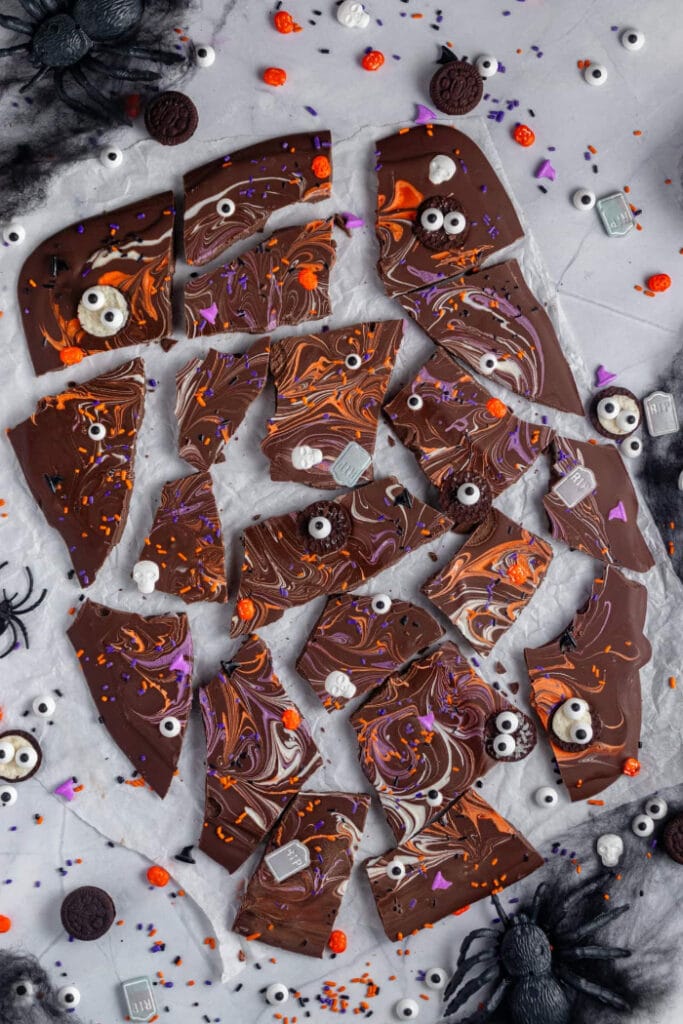 Pieces of chocolate Halloween bark are decorated with orange and purple swirls, candy eyes, sprinkles, mini cookies, skulls, and tiny tombstone candies.