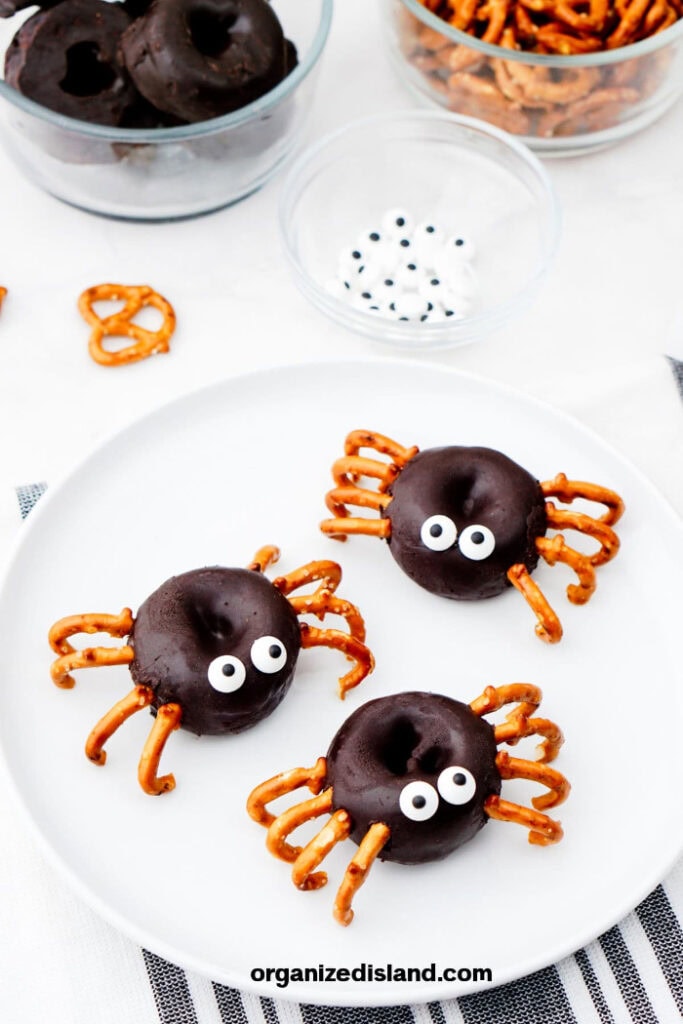 Three chocolate mini donut spiders are decorated with pretzel legs and white candy eyes on a large white plate.