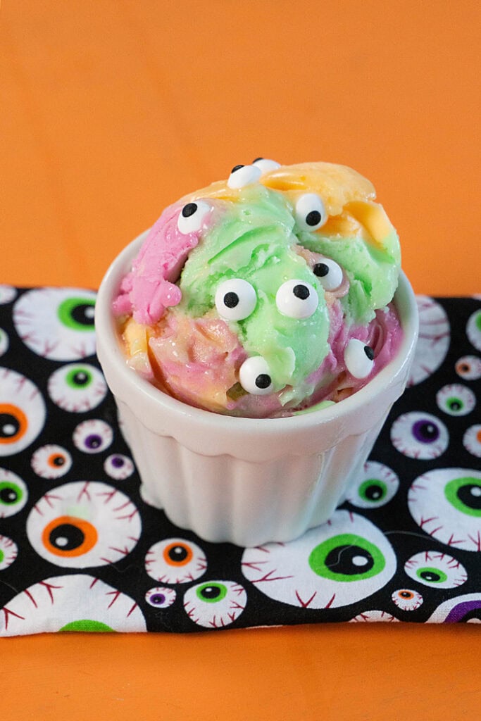 A white ramekin holds colourful pink, green, and orange ice cream topped with several black and white candy eyes on an eyeball patterned napkin.