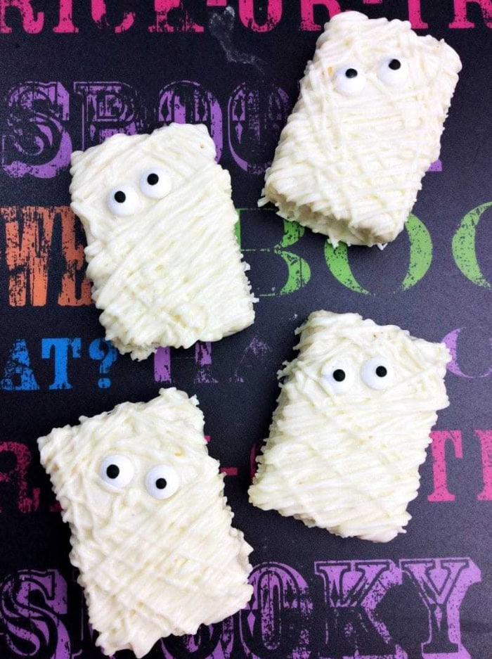 Four rectangular Rice Krispie treats are covered with drizzled white chocolate and decorated with candy eyes to look like playful Halloween mummies.