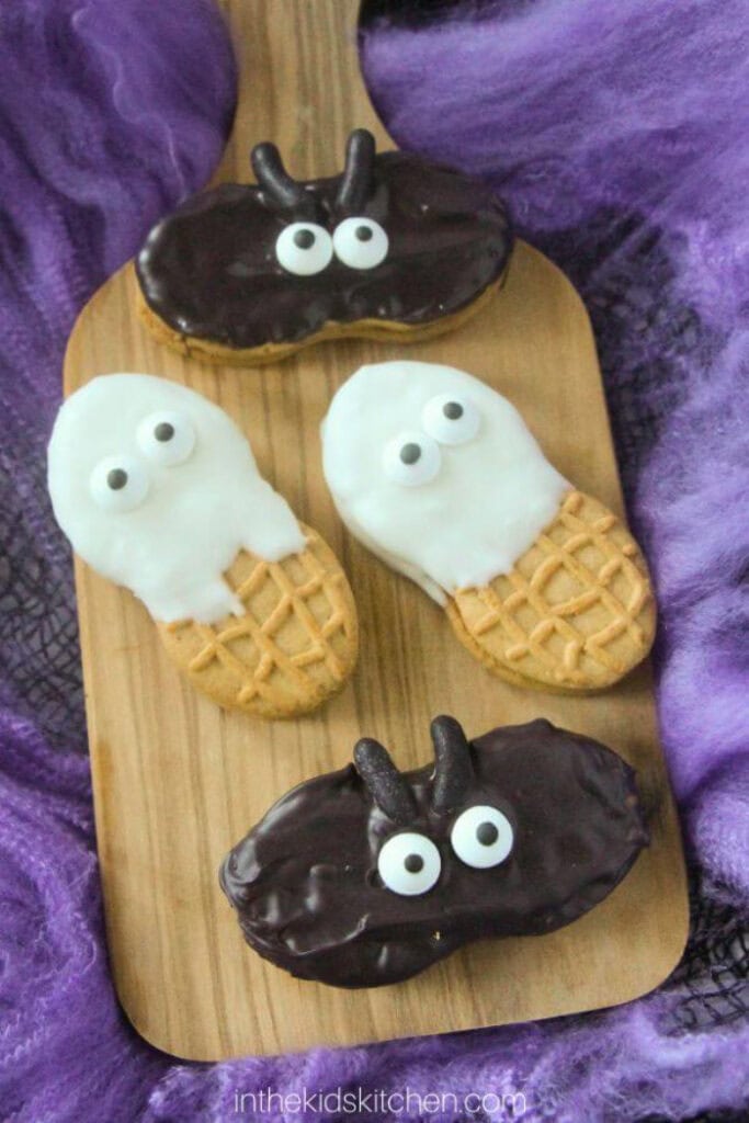 Two white chocolate Nutter Butter ghosts and two chocolate covered Nutter Butter bats are decorated with candy eyes and arranged on a wooden board.